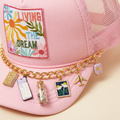 Angled detail view of Living My Dream pink trucker hat and gold travel charm chain on a cream background